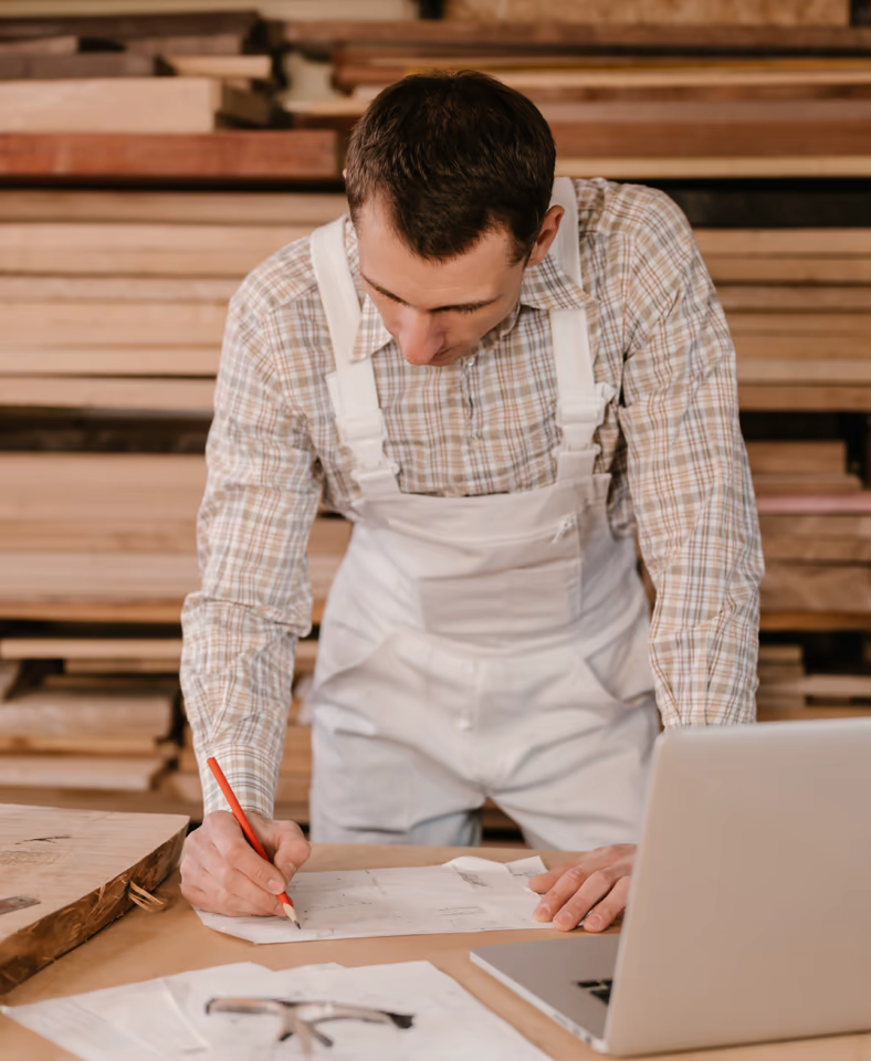 Schreiner in weißer Arbeitsschürze zeichnet Pläne an einem Werktisch in einer Holzwerkstatt