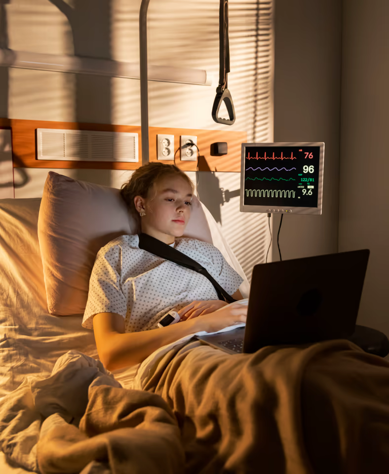 Junge Patientin liegt im Krankenhausbett mit Laptop, neben ihr ein Herzmonitor