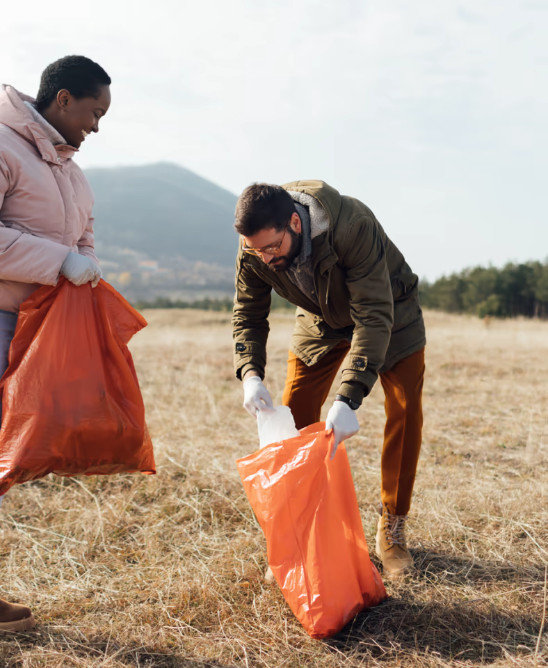 Zwei Personen sammeln mit orangenen Müllsäcken und Handschuhen Abfall auf einem Feld in der Natur
