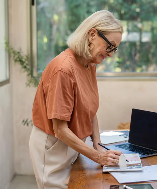 Frau mit Brille arbeitet stehend an einem Schreibtisch mit Laptop und Unterlagen